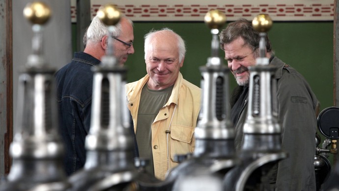 Drei Männer schauen sich die Dampfmaschine an, von der drei Ventile zu sehen sind.